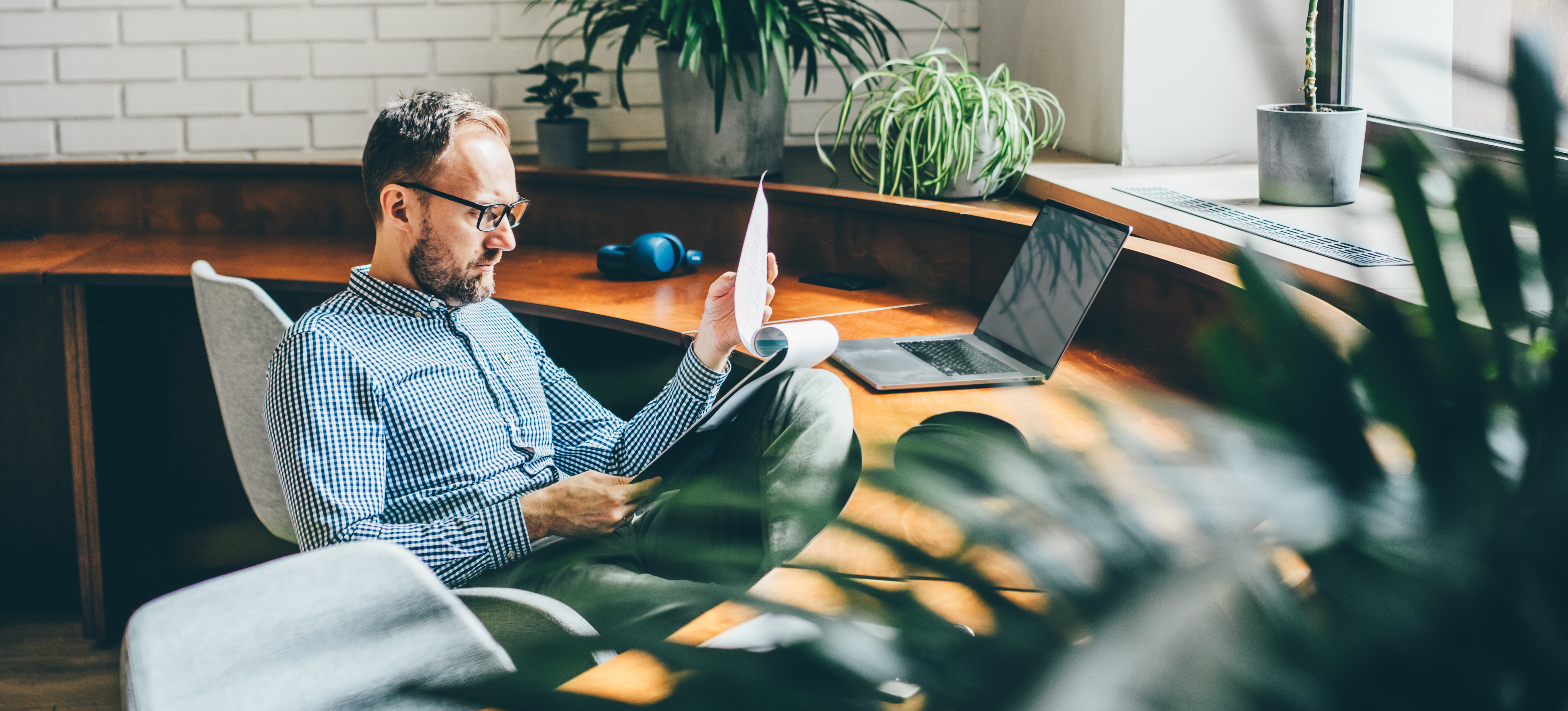 [Featured Image] A stock portfolio manager familiar with stochastic calculus for finance reads over a report as they sit in a pleasant office with multiple potted plants and a sunny window.
