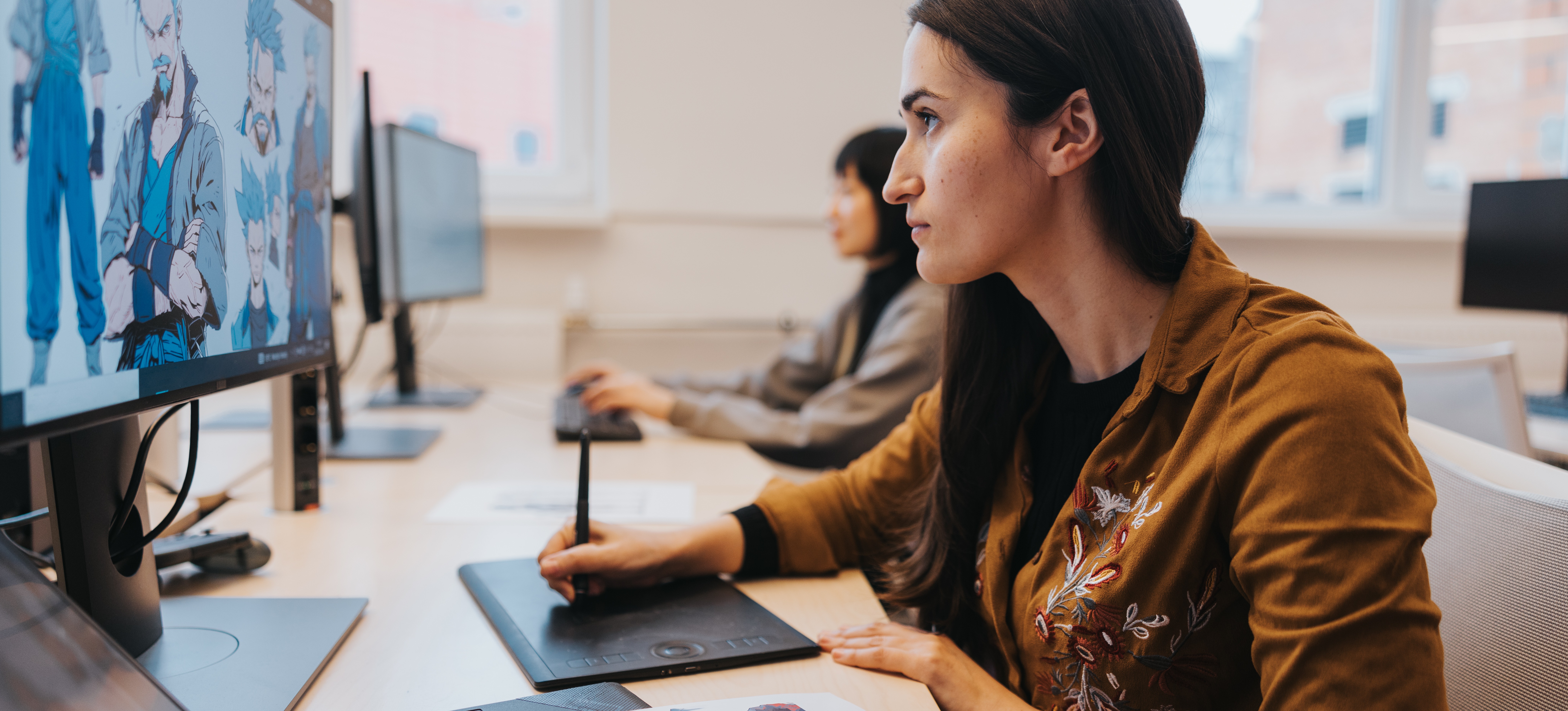 [Featured Image] A video editor sits at her computer next to a colleague and edits frames with her device.
