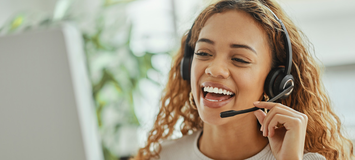 [Featured Image] A smiling woman IT support specialist talks on a headset to a customer as she looks at a desktop in front of her.
