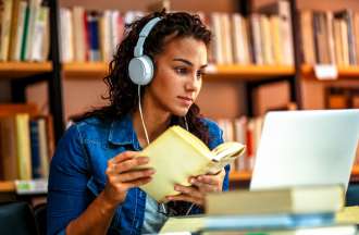[Featured Image] A college student wears headphones and sits at a library table with a book in her hands and a laptop in front of her.