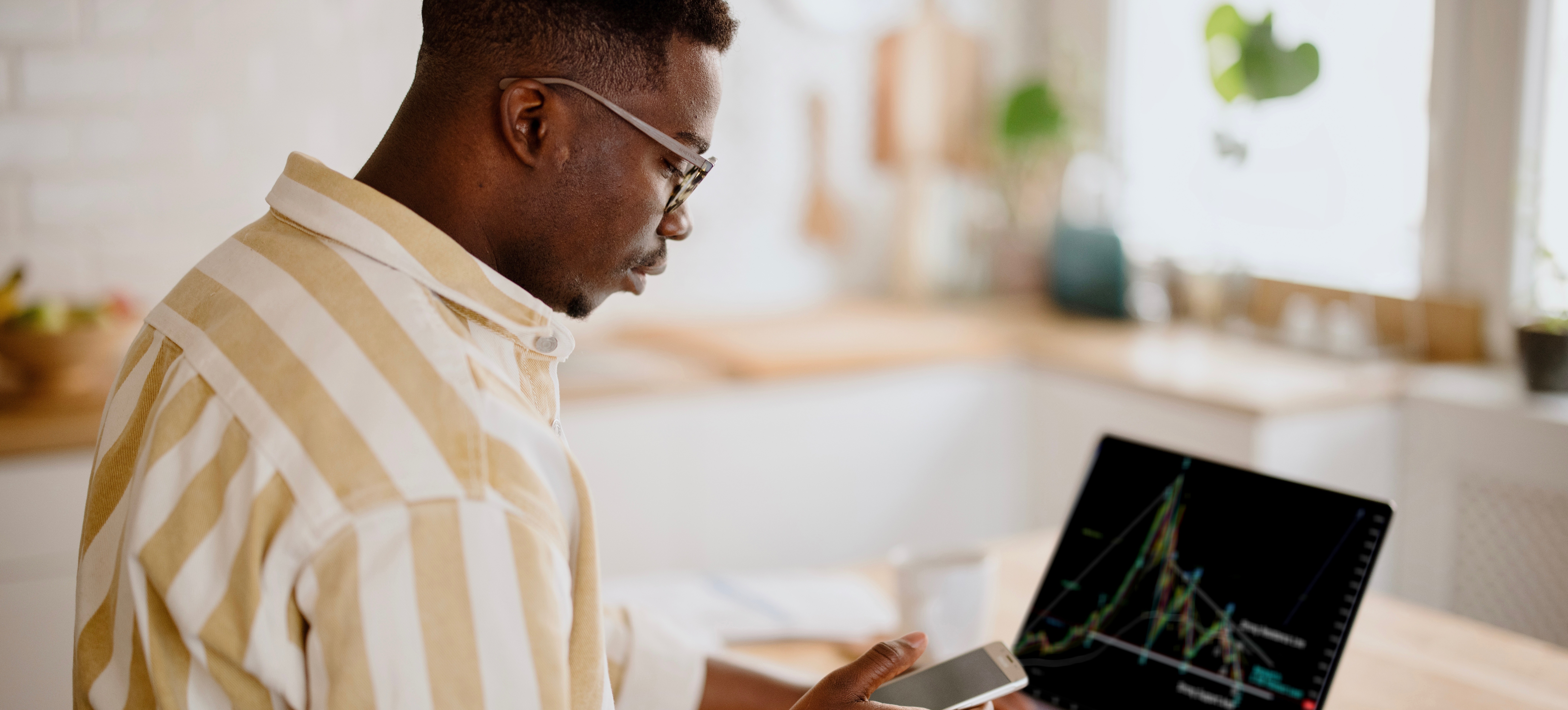 [Featured Image] After learning what is crypto mining, a man sitting in his kitchen watches his new crypto investments on his open laptop and his phone simultaneously.
