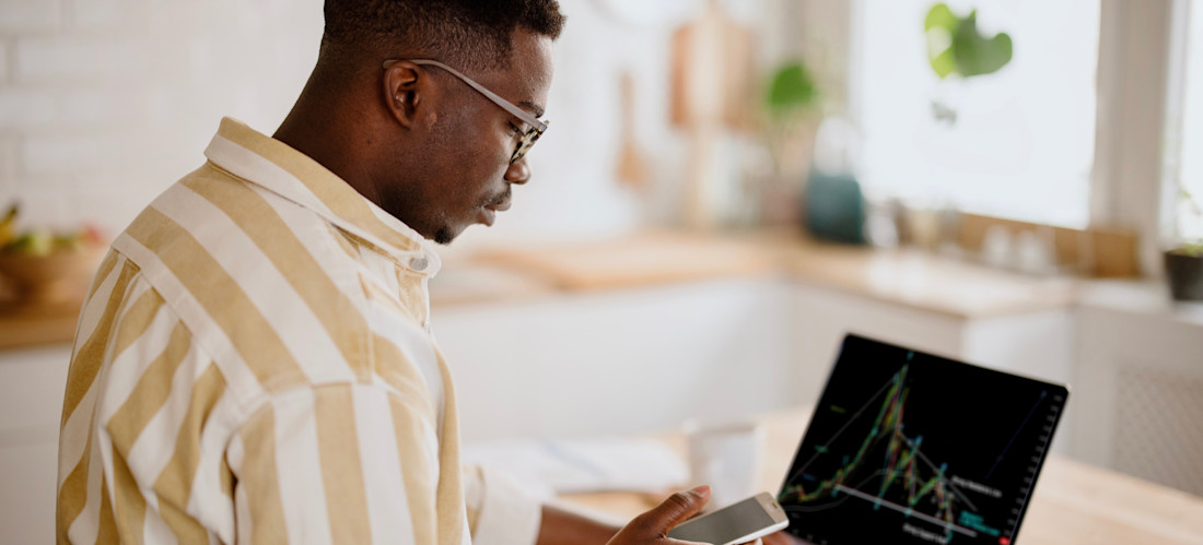 [Featured Image] After learning what is crypto mining, a person sitting in their kitchen watches their new crypto investments on their open laptop and their phone simultaneously.
