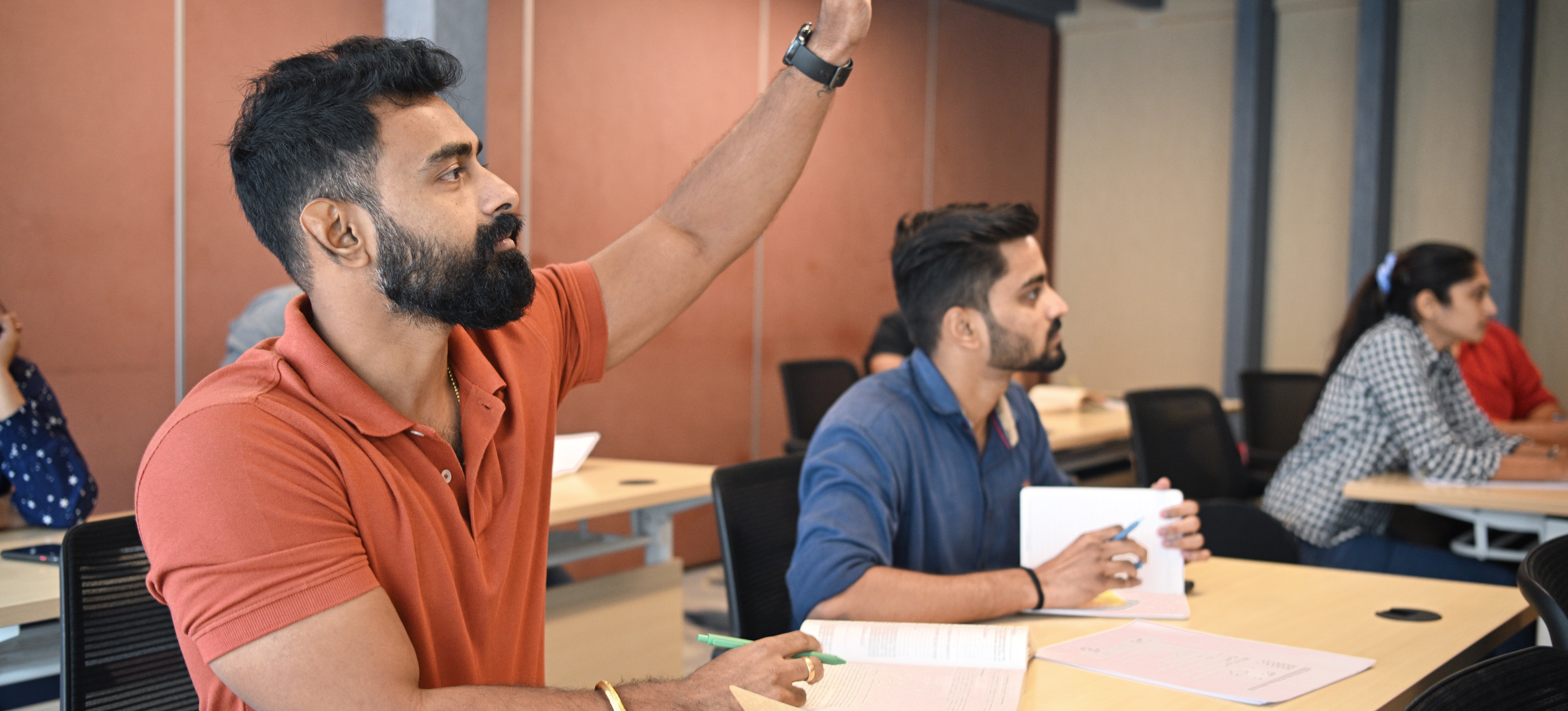 [Featured Image]: A learner in a classroom raises their hand to ask a question as they take a class as part of an upskilling plan.
