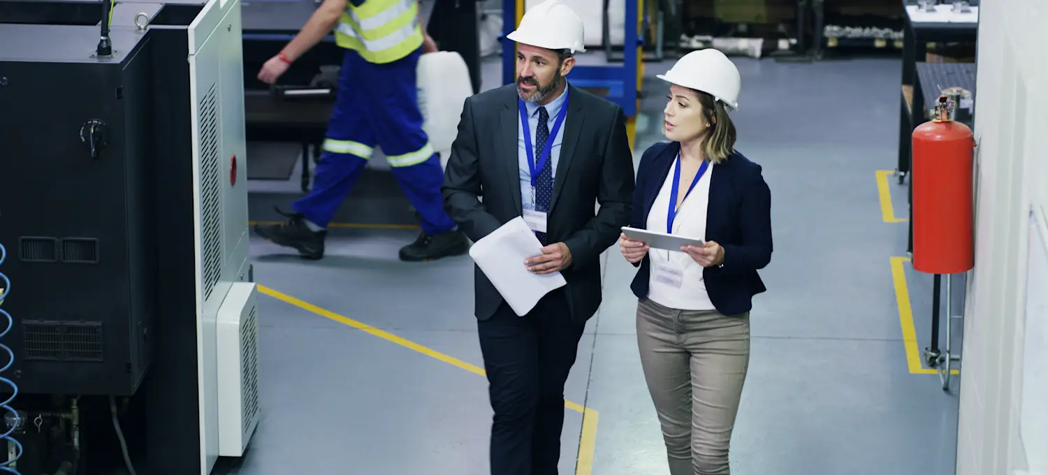 [Featured Image] Two people, wearing hard hats and both working for an operations engineer salary, walk through a factory and discuss how to improve the factory's industrial systems and processes.
