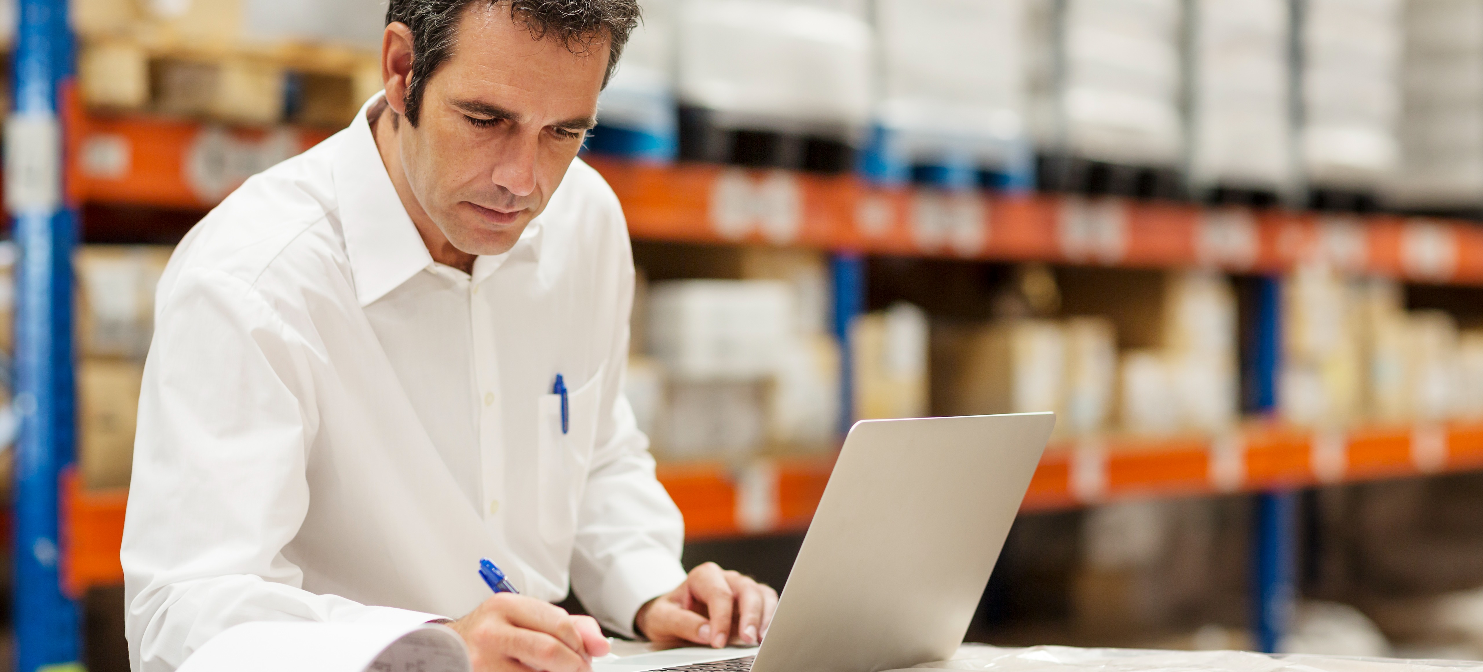 [Featured Image] A supply chain professional is conducting inventory management in a warehouse as they write with a pen on an inventory sheet and makes calculations on their laptop.