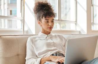 [Featured Image} A woman interested in an IT career relaxes on a sofa and searches on her laptop, "What is the easiest IT certificate to get."