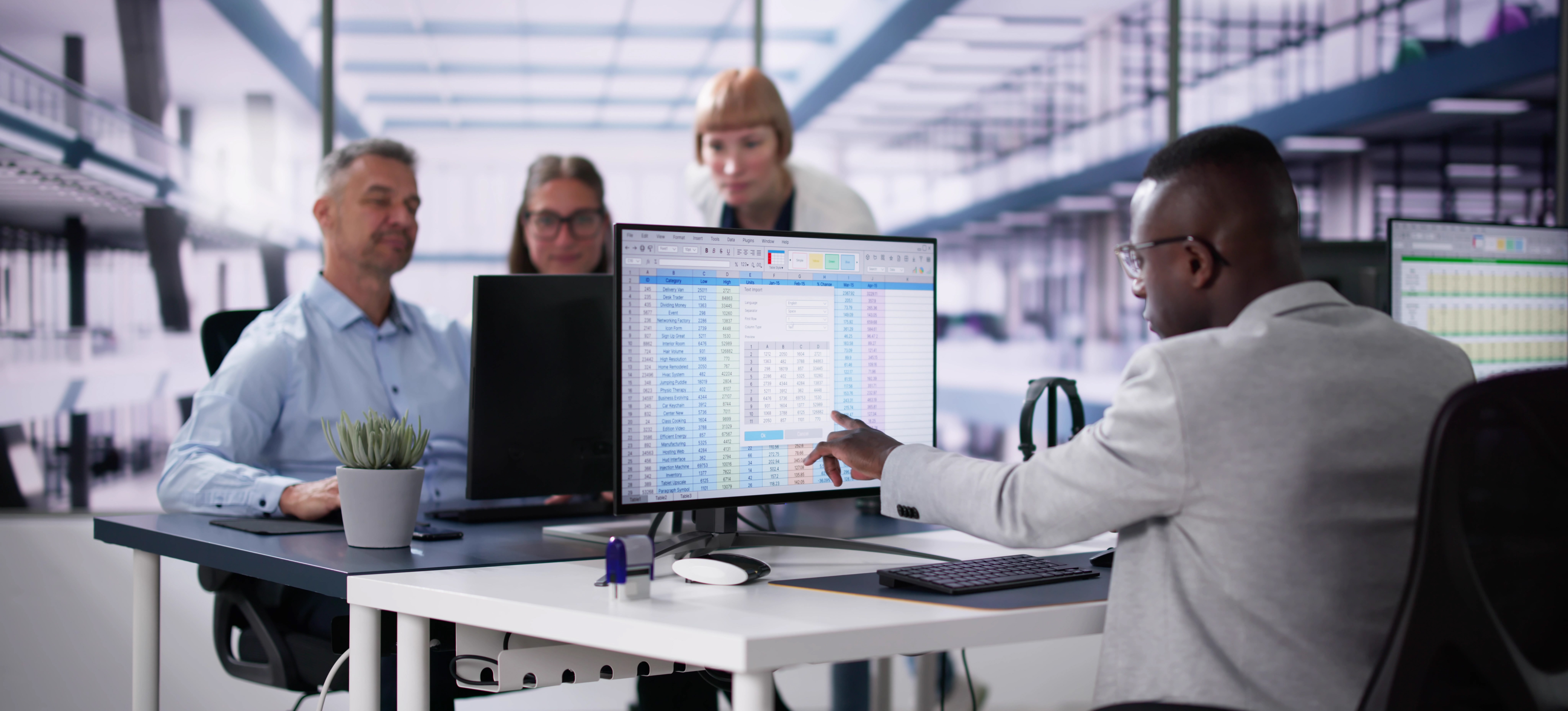 [Featured Image] A person in a business suit uses Excel structured references as they point to a spreadsheet on their computer monitor, and their teammates look at another screen.
