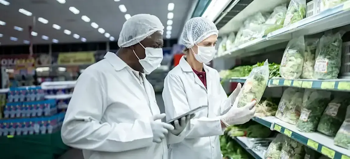 [Featured Image] Two health inspectors dressed in white lab coats, gloves, hair nets, and face masks hold clipboards as they inspect packages of vegetables in a supermarket.

