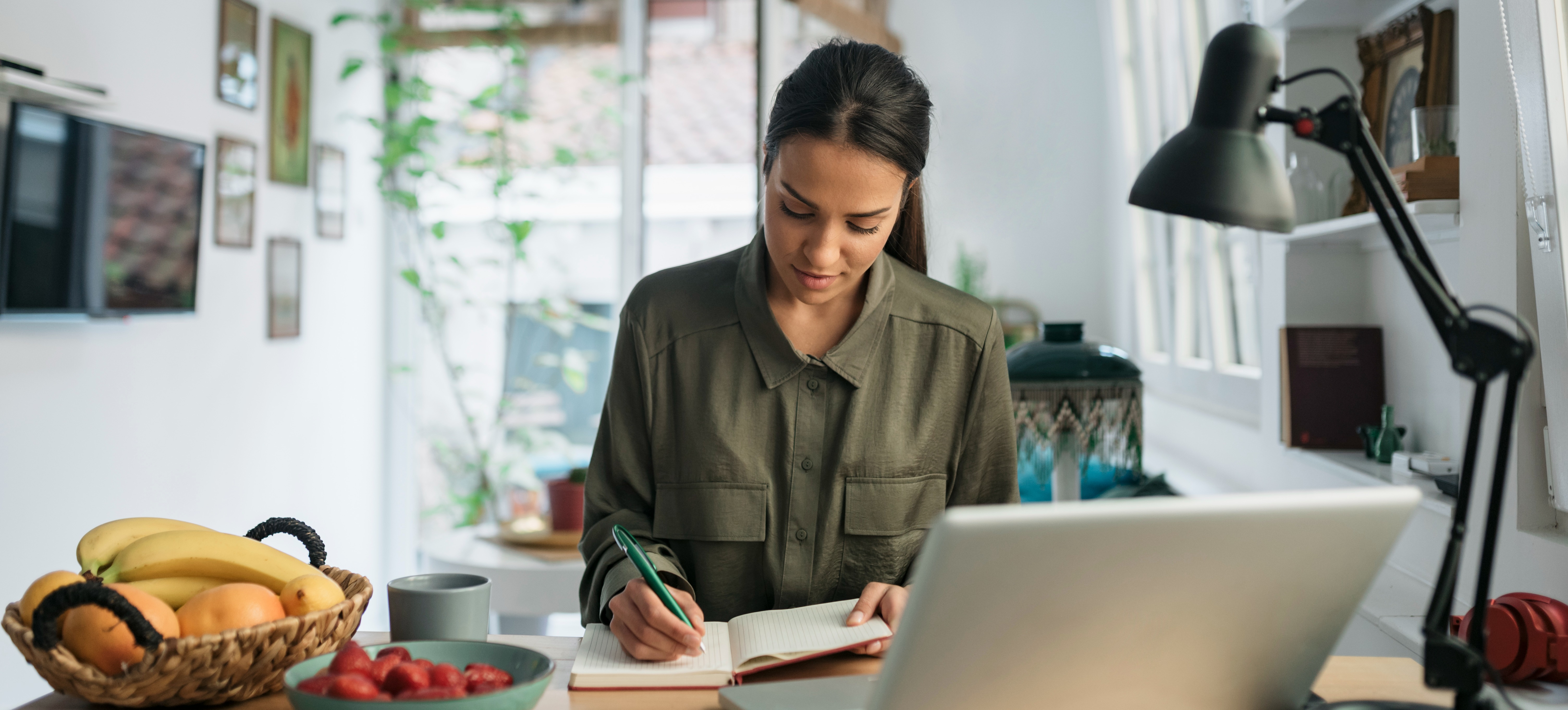 [Featured image] Person wearing green top sits across a laptop and writes down steps to add columns in Google Sheets for future reference.