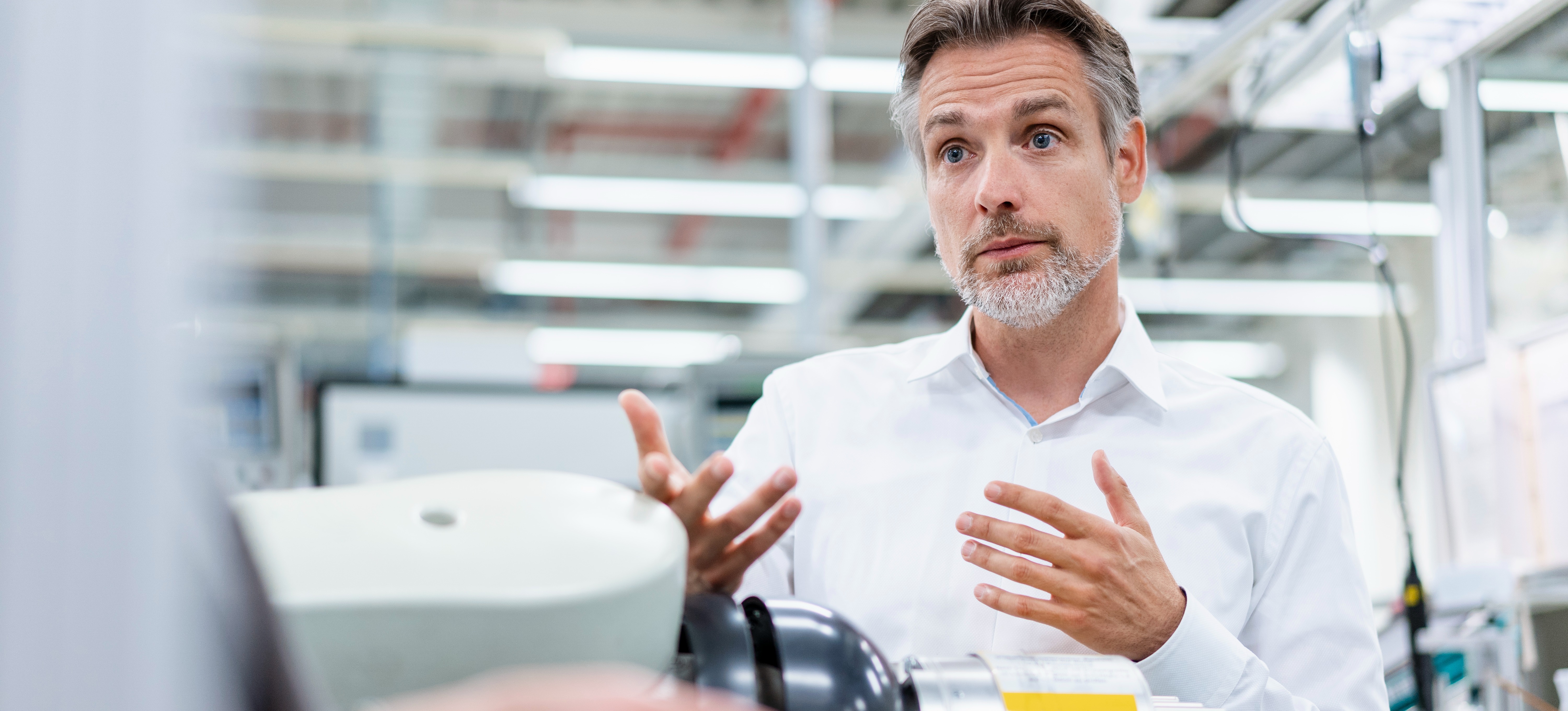 [Featured Image] A businessman is talking to a colleague about responsible AI next to an assembly robot in a factory.
