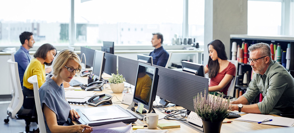 [Featured Image] Six employees work at their computers and workstations thanks to an effective network switch that allows them to connect to the local area network seamlessly.
