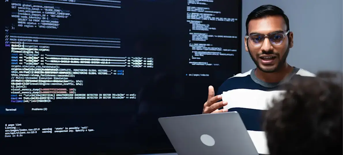 [Featured Image]: A person stands in front of a large monitor and holds a laptop in their hands as they speak about concepts they learned for a DevOps degree.