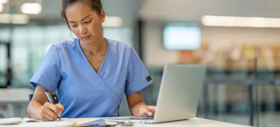 [Featured Image] A nursing student wearing scrubs uses a pen and laptop to do their coursework.  