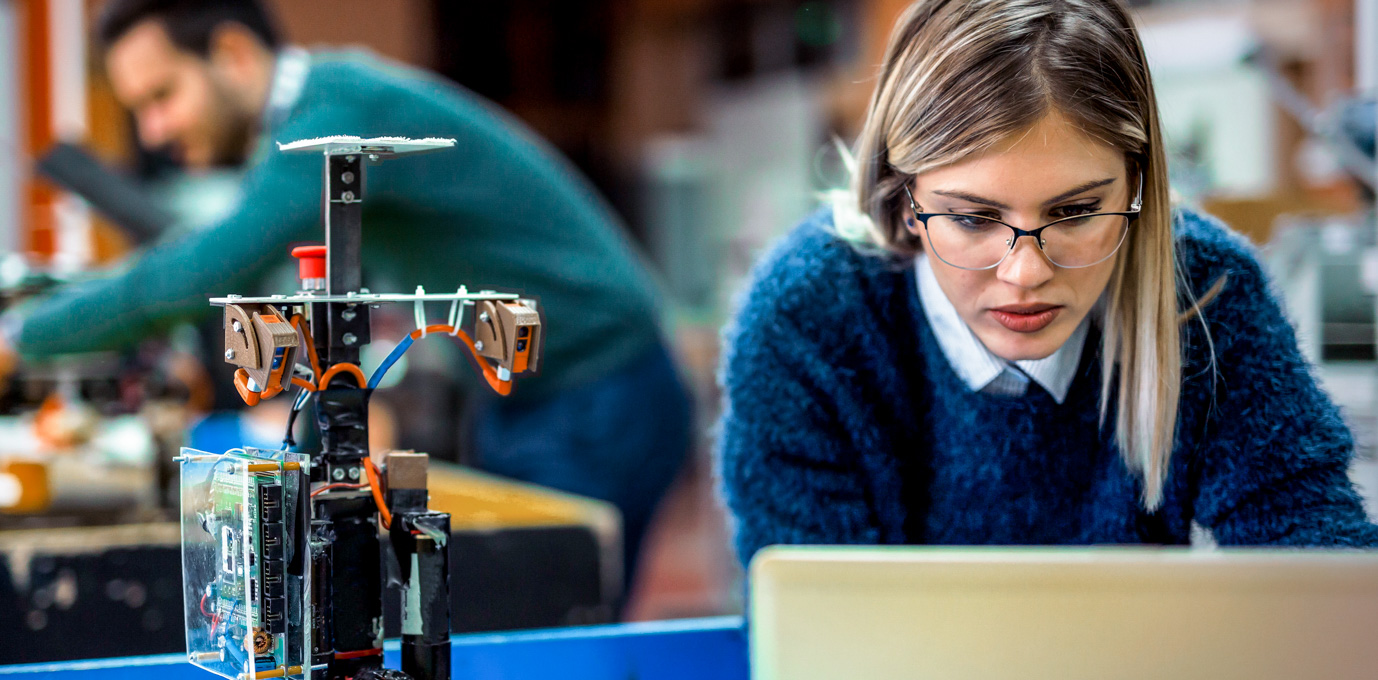 [Hauptbild] Eine Person mit Brille und blauem Pullover arbeitet vor einem Computer in einem futuristischen Informatiklabor