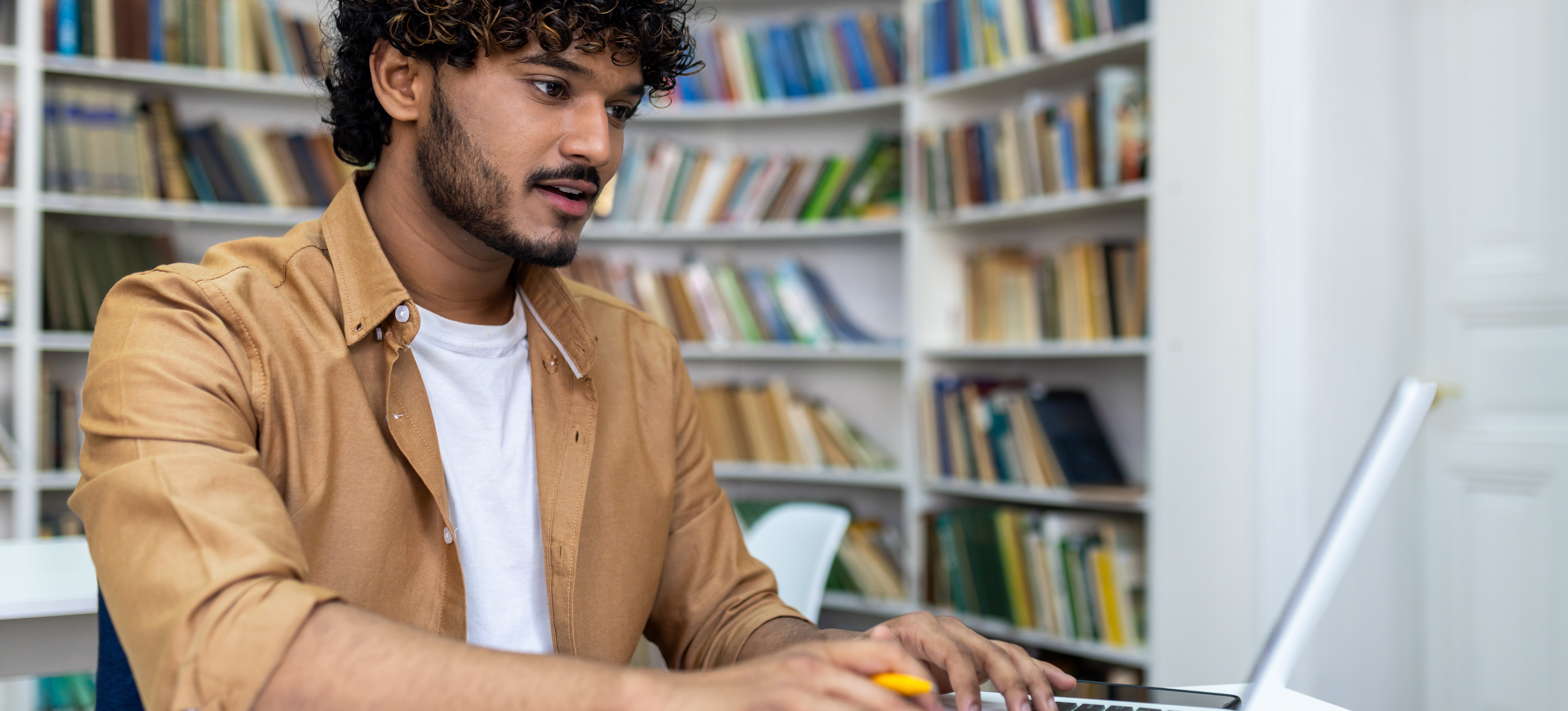 [Featured Image] A postgraduate learner in a brown button-down shirt over a white T-shirt studies on their laptop in a library as they work toward their master’s in data science.
