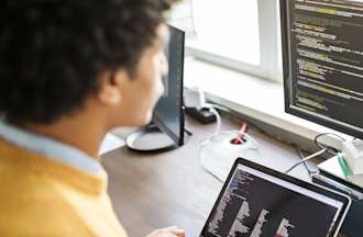 [Featured Image]:  A male, wearing a yellow jacket, is sitting at his desk, working on his desktop and laptop, performing his duties in his data visualization job. 