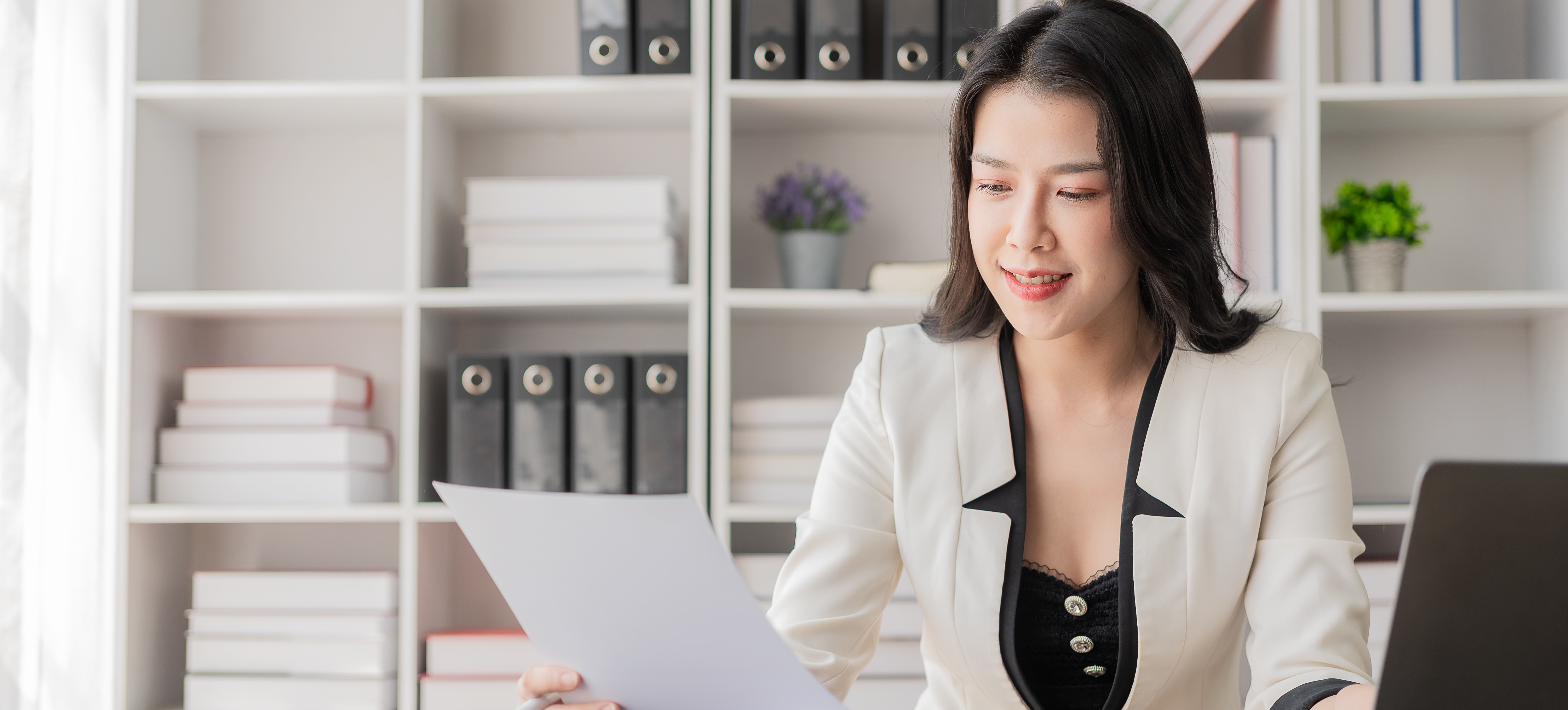 [Featured Image] A businessperson in a white blazer and black top with gold buttons smiles while reviewing an accounts payable report in an office with white shelves, black binders, and books. 
