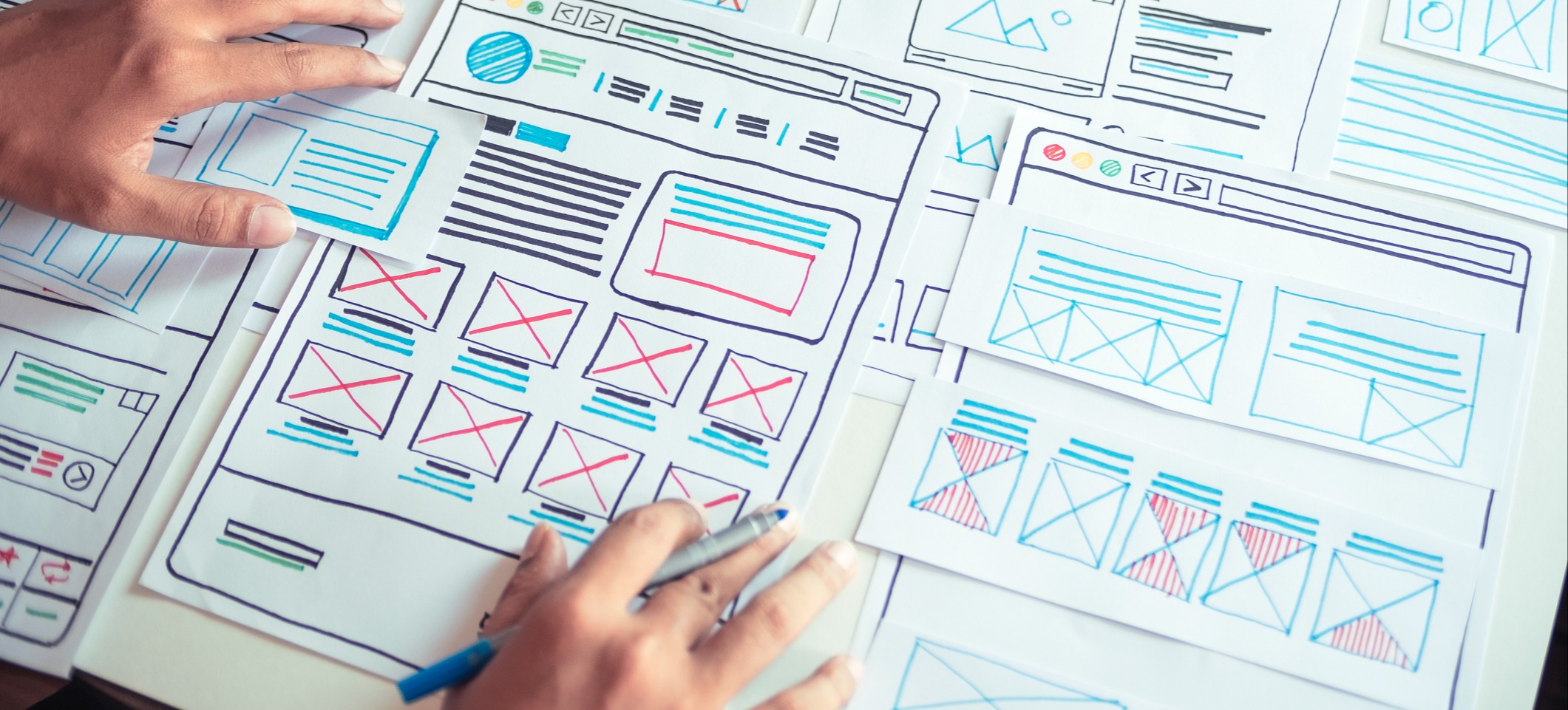 [Featured Image] A UX designer utilizes negative spaces as he hand draws wireframes with colored markers on sheets of paper at a desk.
