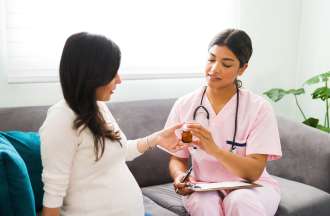 [Featured Image]:  A female, nurse midwife, wearing a pink uniform, and a stethoscope around her neck, is talking to a pregnant woman, wearing a white top and black pants.  They are sitting on a couch,, with two blue pillows. with a plant on one side,