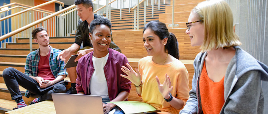 [Featured Image] Three business school students sit on wooden steps in the foreground, with two more students sitting in the background, chatting and smiling in two separate groups as they refer to laptops and books in front of them.