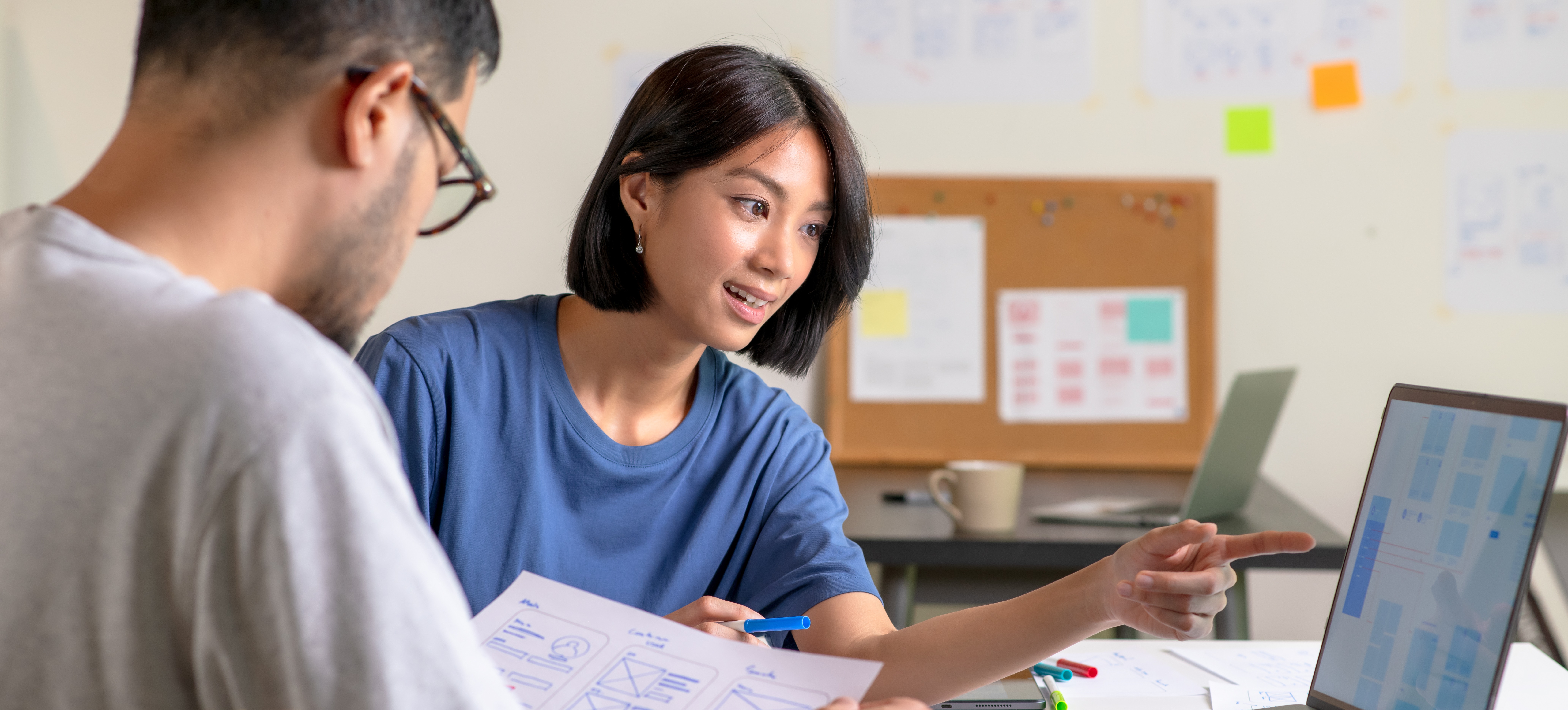 [Featured Image] UX designers discussing wireframes for a website and mobile app prototype. One designer is looking at a low-fidelity sketch of the website, while another is looking at a laptop.
