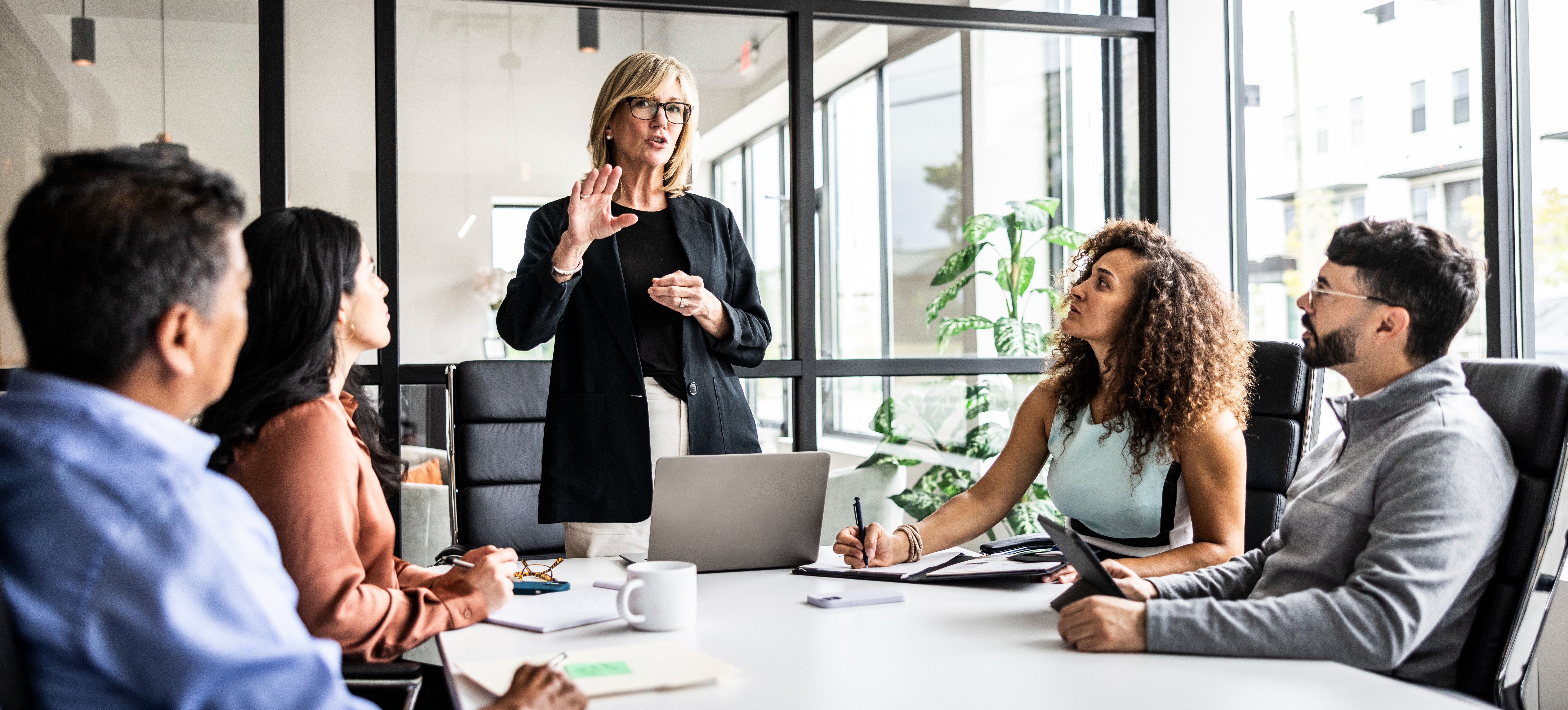 [Featured Image] A female manager stands and talks to a group of her employees in an office and shows leadership skills.