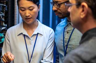 [Featured Image] Three mainframe developers looking at a digital tablet and talking while standing next to a server in a data center.