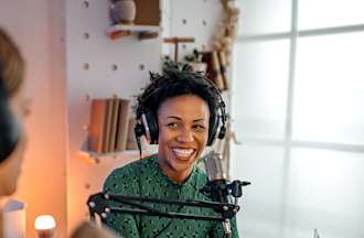 [Featured image] Two females, one wearing a green sweater and the second with long brown hair, are sitting in a room with a microphone wearing headphones on a desk, as they prepare to broadcast their cybersecurity podcast.