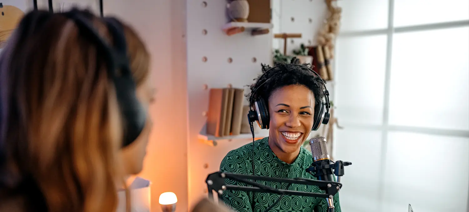[Featured image] Two females, one wearing a green sweater and the second with long brown hair, are sitting in a room with a microphone wearing headphones on a desk, as they prepare to broadcast their cybersecurity podcast.