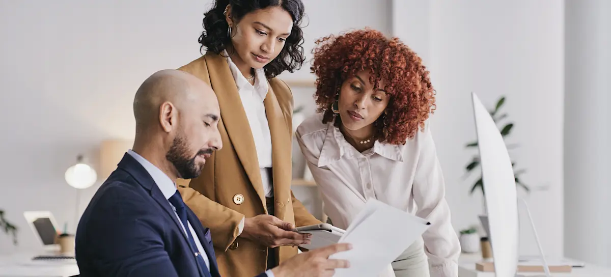 [Featured Image] Three members of a business’s human resources team discuss the business systems analyst salary range for a new hire.

