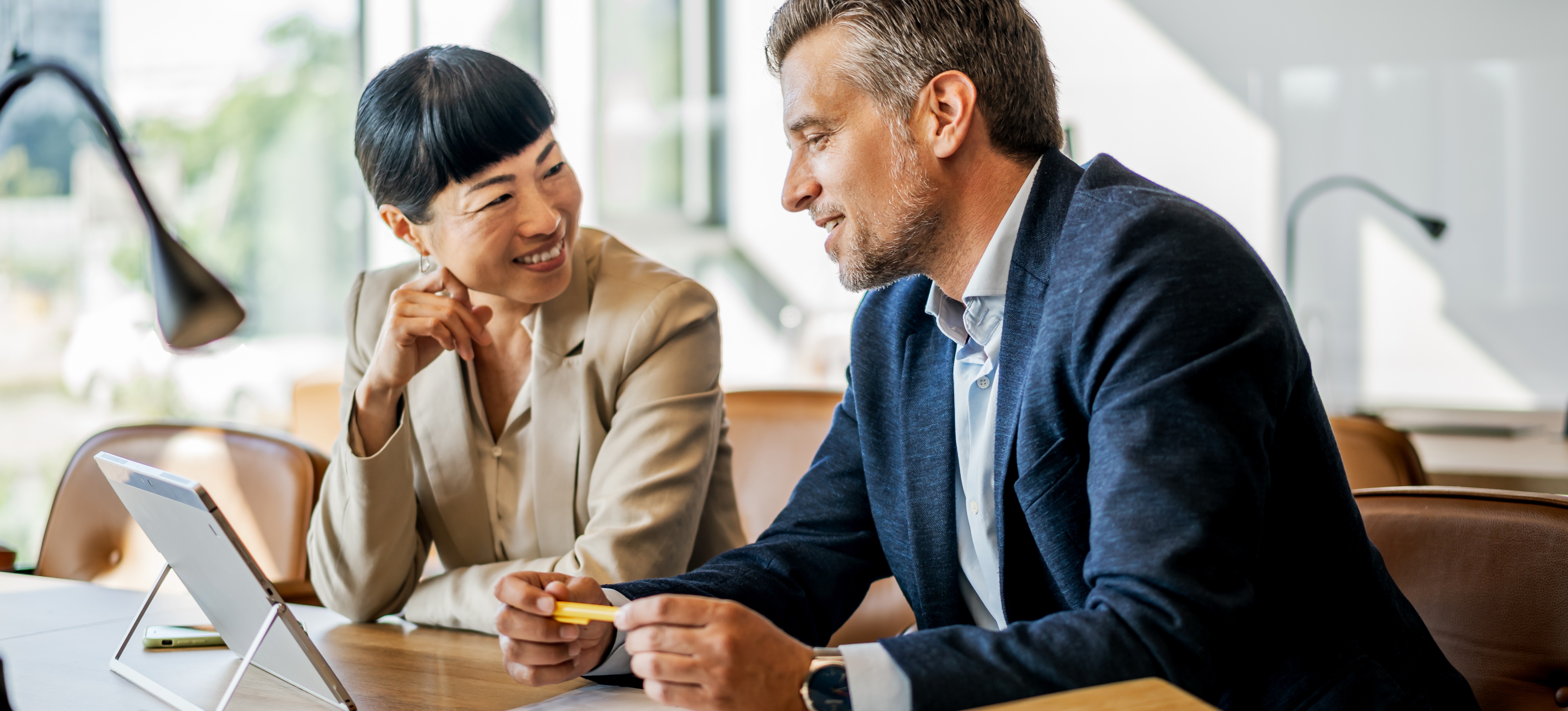 [Featured image] Two financial professionals are performing an income analysis. They are  sitting side by side at a desk, looking at an income statement on a tablet. They are both smiling. There is a window with a view of the outdoors in the background.