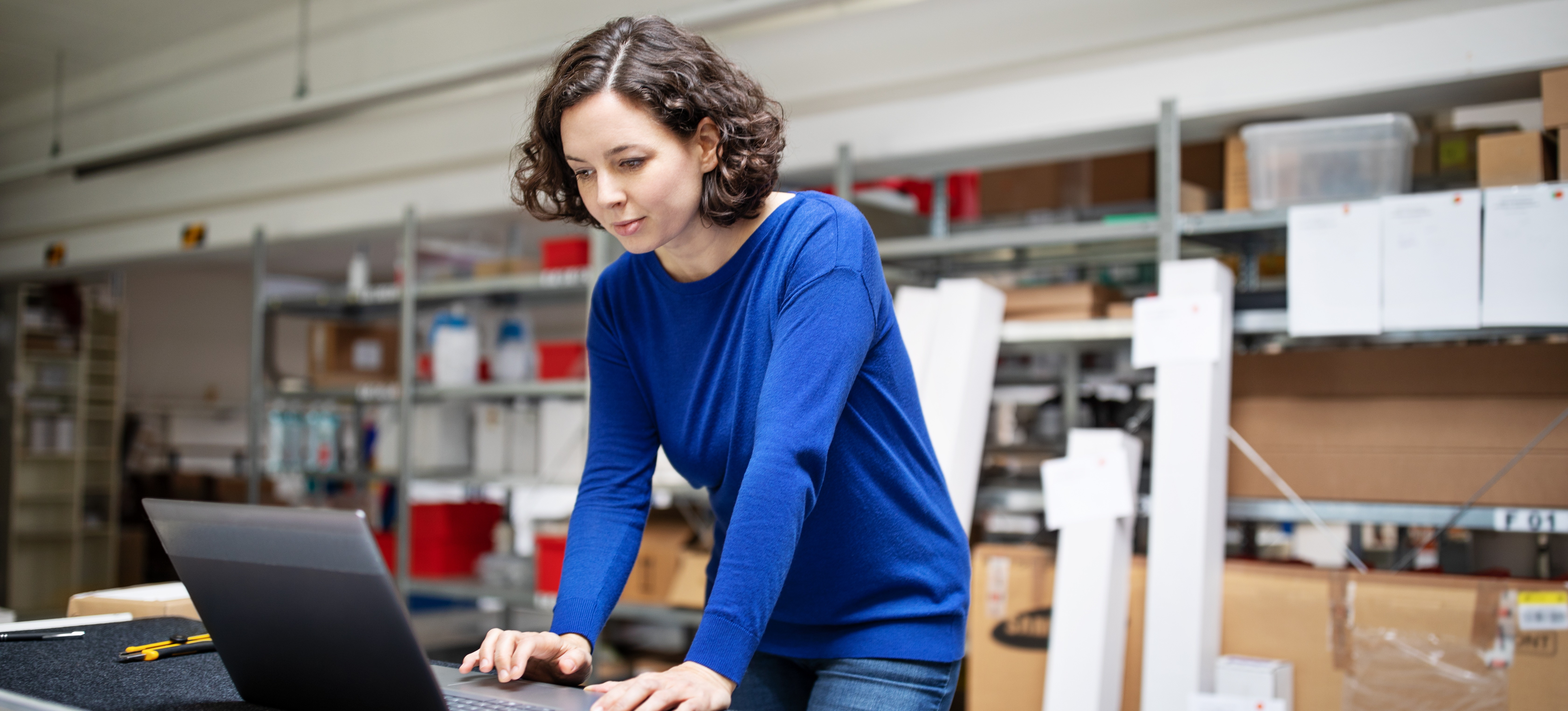 [Featured Image] A supply chain analyst in a blue shirt and denim jeans stands at a table in a supply room, working on her laptop.
