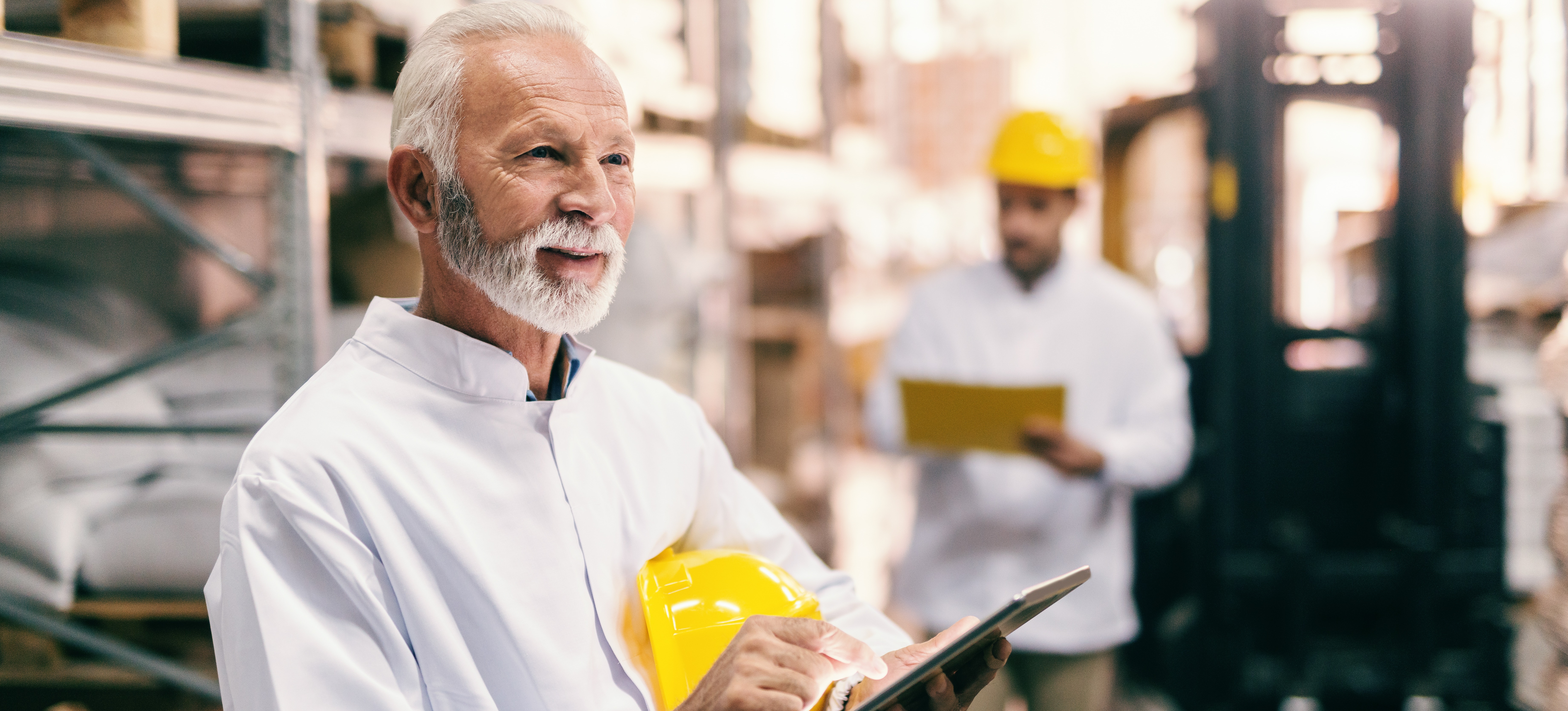 [Featured Image] A person with supply chain certification walks around a warehouse with a tablet, while their coworker waits in the background with a forklift.
