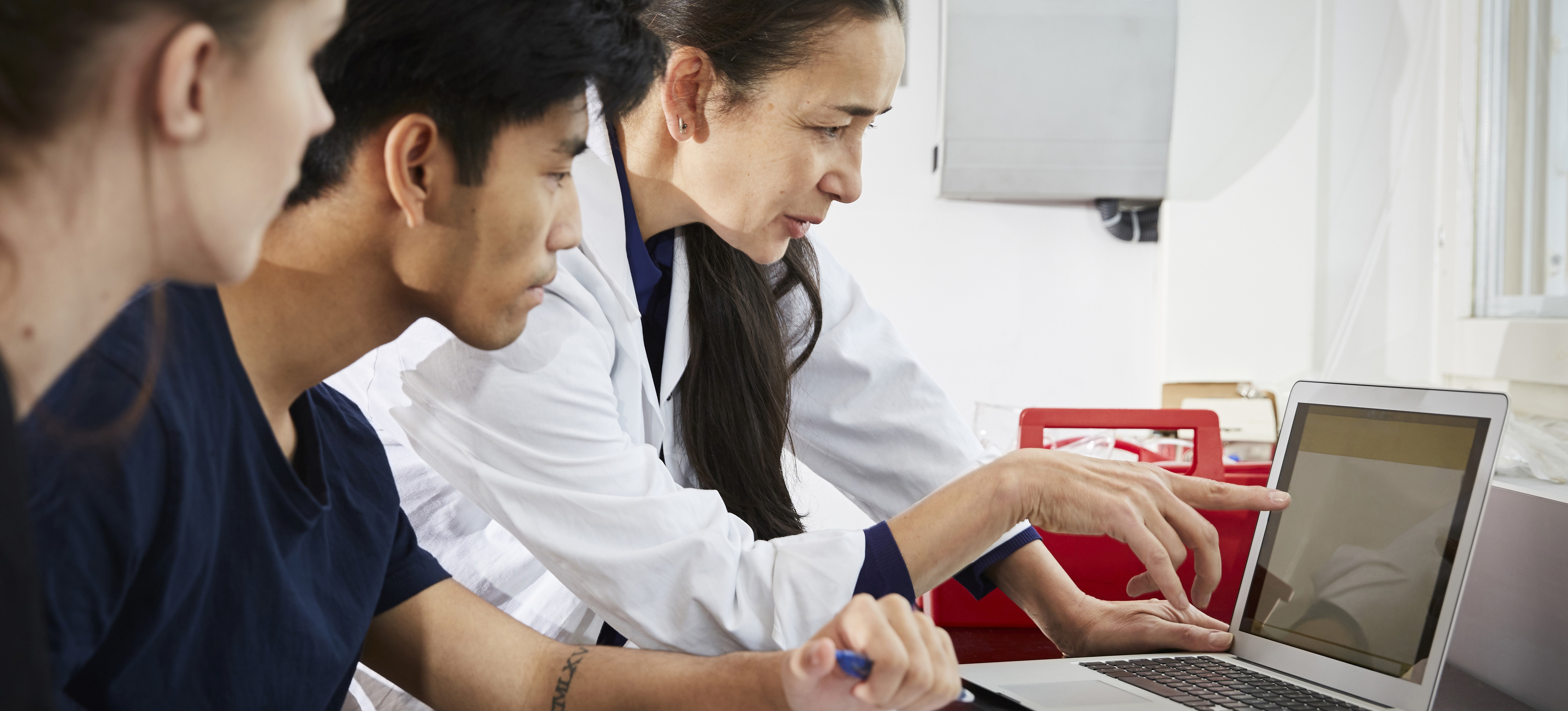 [Featured image] Three people look at a computer. One, who is a certified pharmacy technician, has a lab coat on.