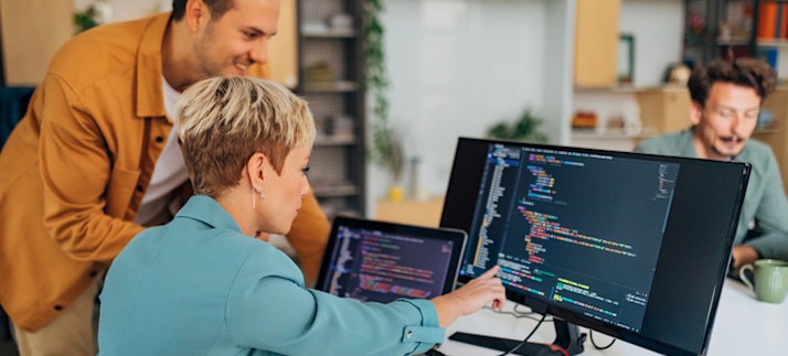 [Featured Image] Two colleagues in an open-space office look at a computer monitor displaying HTML and discuss using CSS strikethrough.
