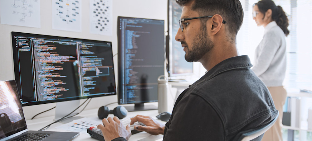 [Featured Image] Two colleagues study code on large computer screens, using cloud security tools to help protect their organization’s cloud data.
