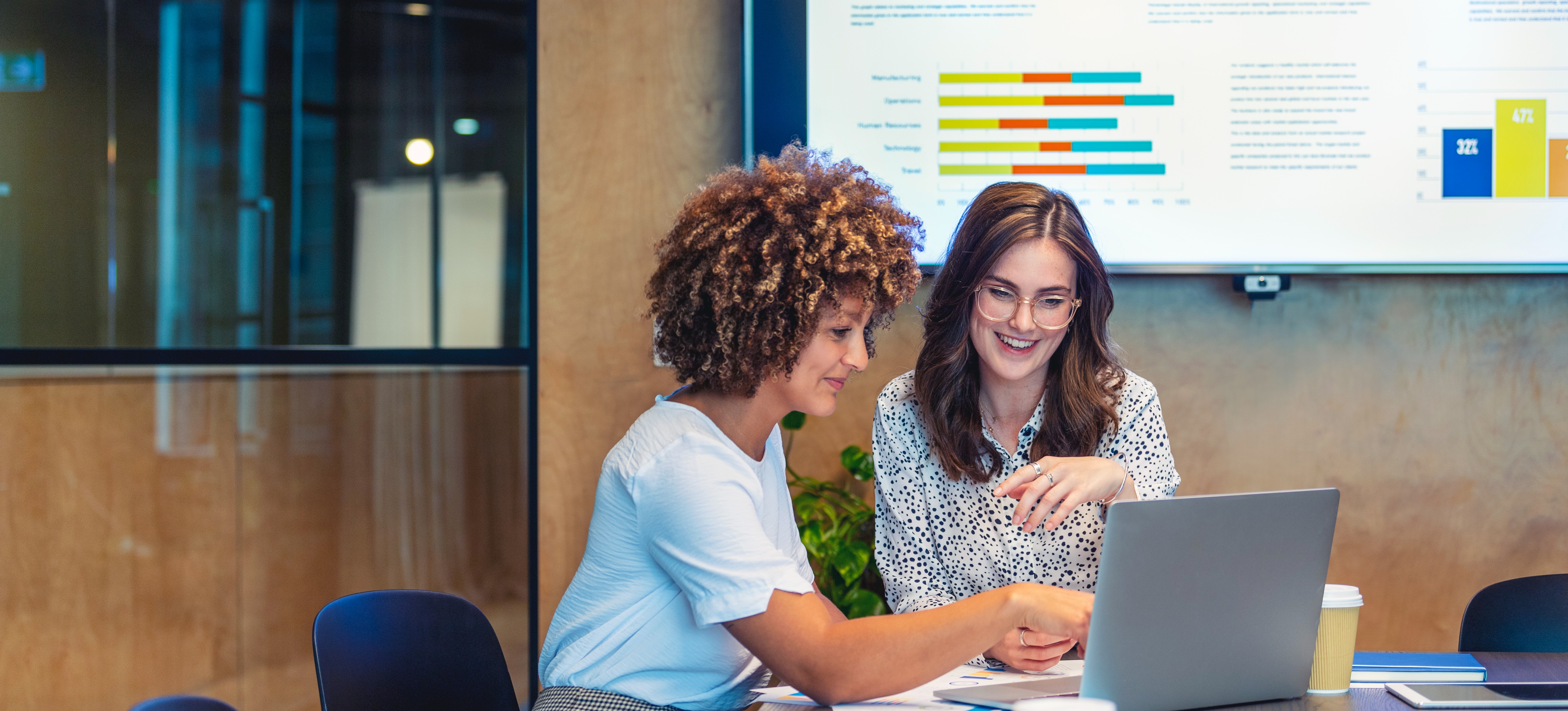 [Featured Image] Two data analysts confer about data analysis methods in front of a laptop while sitting in a boardroom that has a large display of various types of graphs on a screen in the background.
