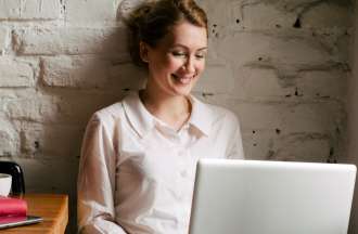 [Featured image] A virtual assistant wears a white blouse and works remotely on their laptop with a red planner and coffee cup to their right.