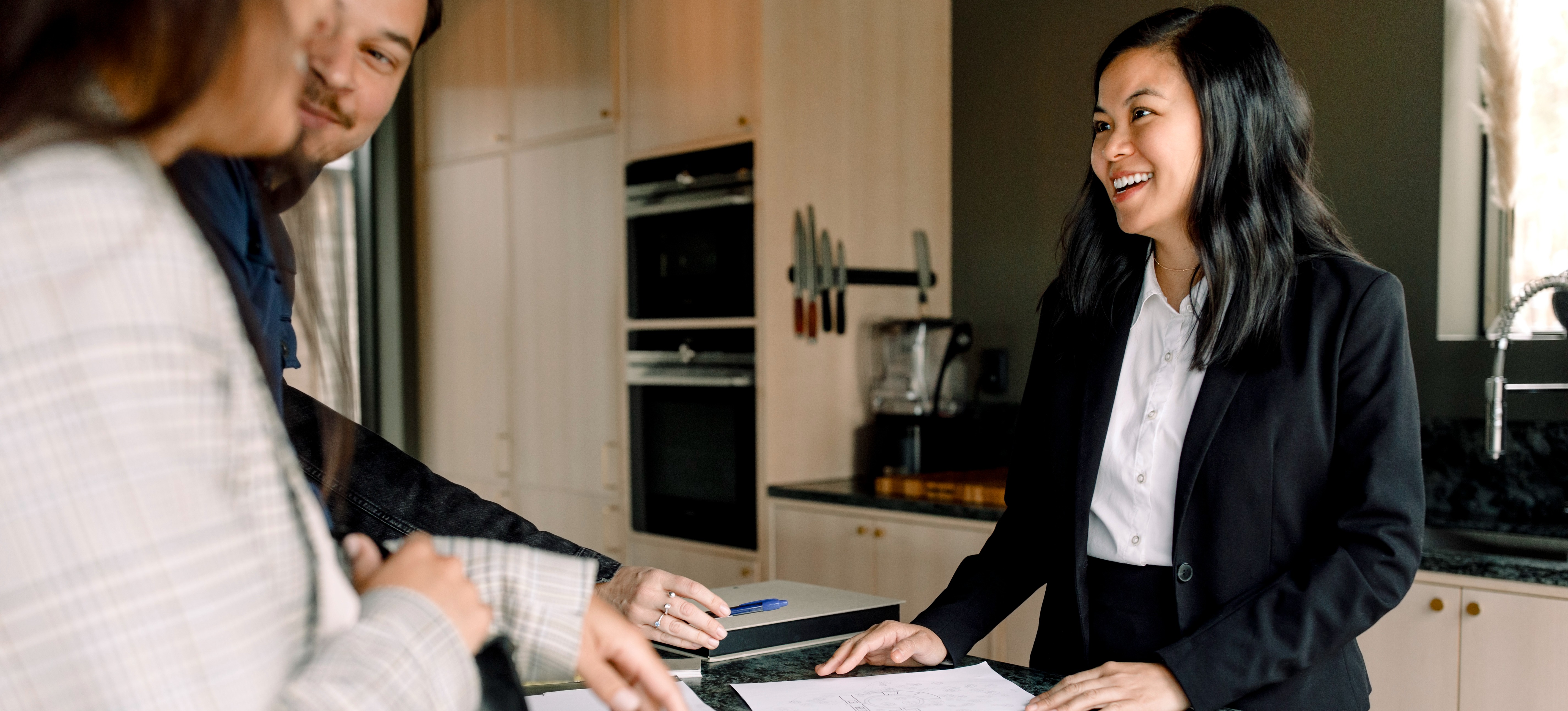 [FEATURED IMAGE] A Realtor stands in the kitchen of a home with paperwork for two people interested in purchasing a house.

