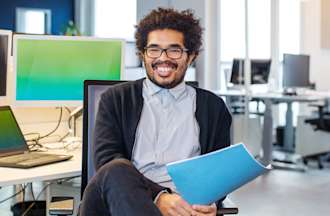 [Featured image] Business Analyst holds file while sitting in front of a laptop computer and monitor