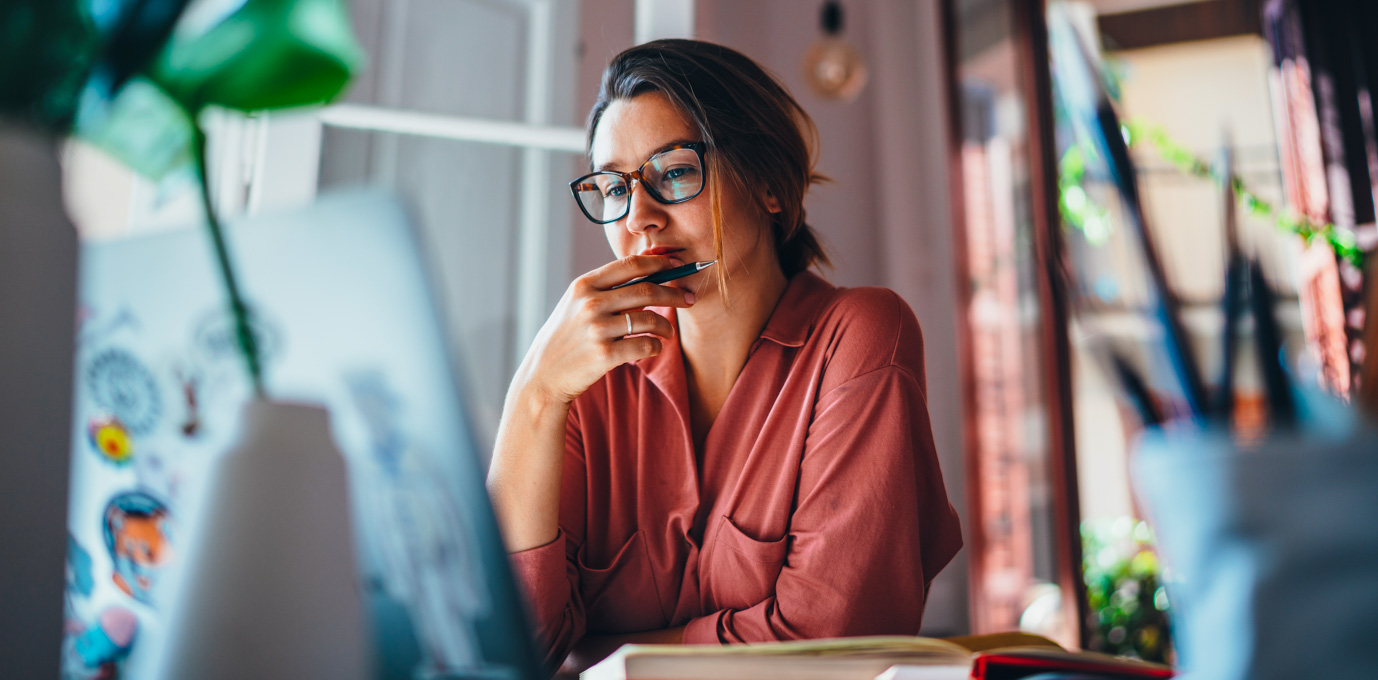[Image en vedette] Un étudiant en informatique vêtu d'un chemisier rose est assis à un bureau avec un ordinateur portable et un livre et réfléchit à la certification informatique d'entrée de gamme à obtenir.
