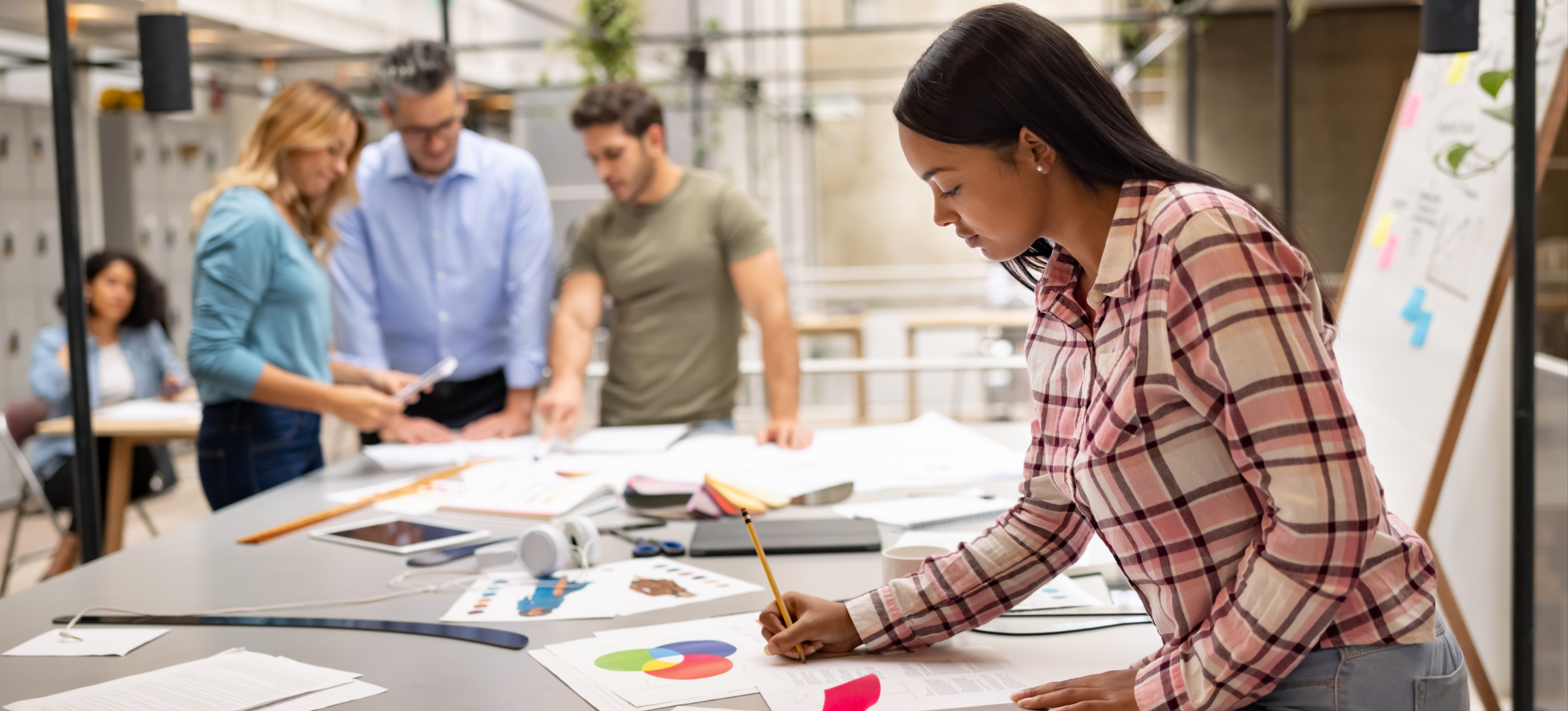 [Featured Image] A designer is working on a sketch at a creative office while a team is brainstorming ideas for a project.