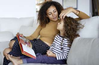 [Featured image] A stay-at-home mom with curly brown hair sits next to a young girl, reading a storybook. 