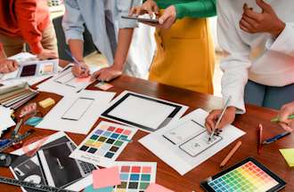 [Featured image] A group of digital designers reviews printouts of color schemes on a large table for mobile and tablet designs.