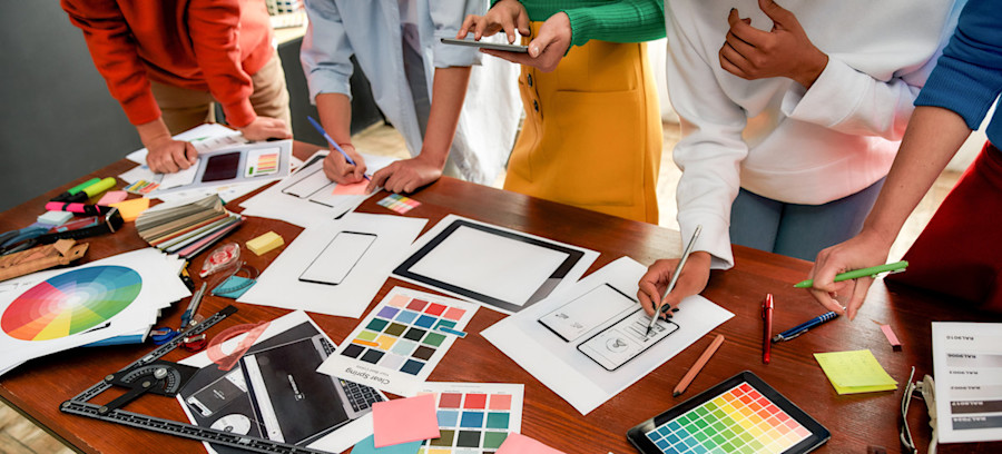 [Featured image] A group of digital designers reviews printouts of color schemes on a large table for mobile and tablet designs.