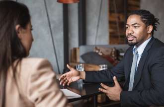 [Featured image] A young Black man in a business suit holds a conversation with a white woman in a business suit. 