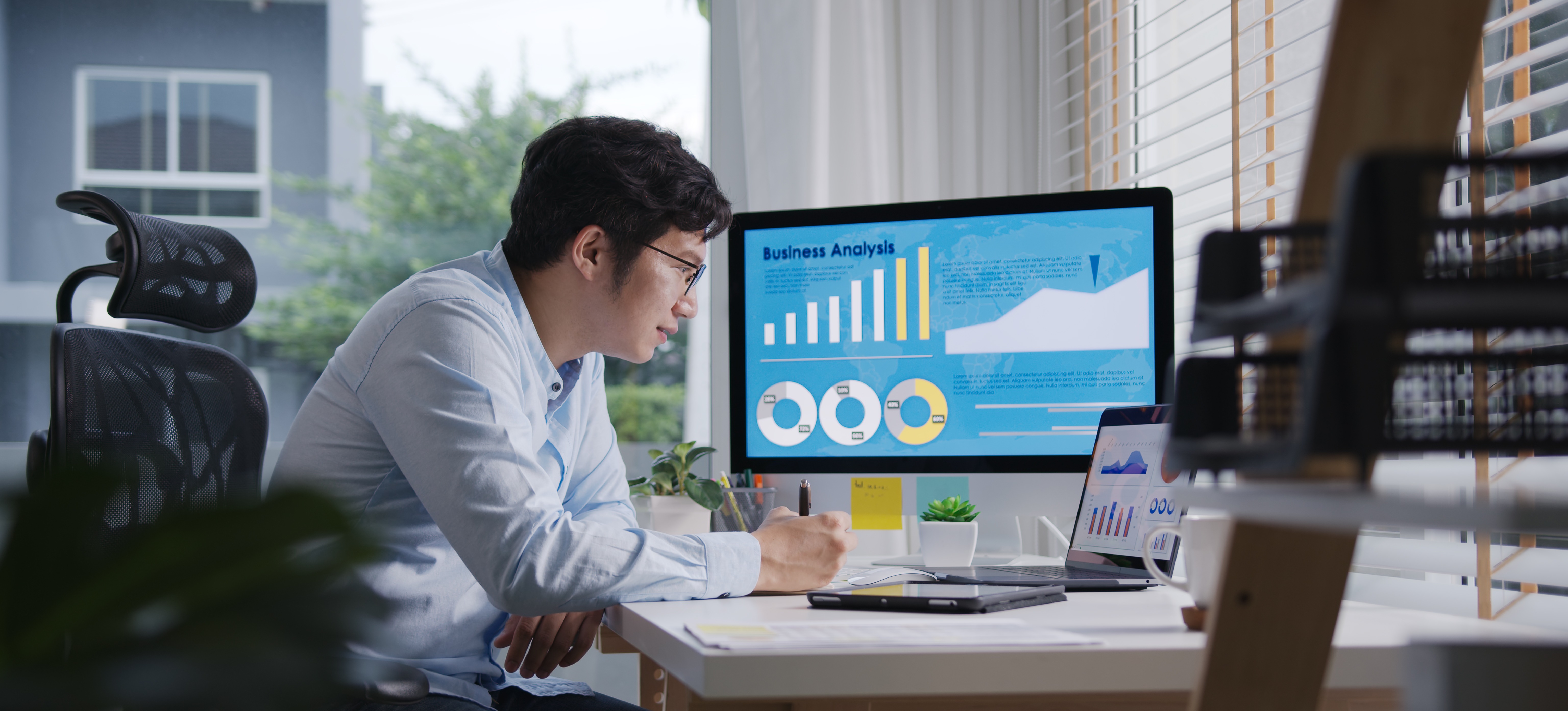 [Featured Image] A businessman sitting at his sunny home office uses KPI dashboards displayed on his computer to analyze his business.
