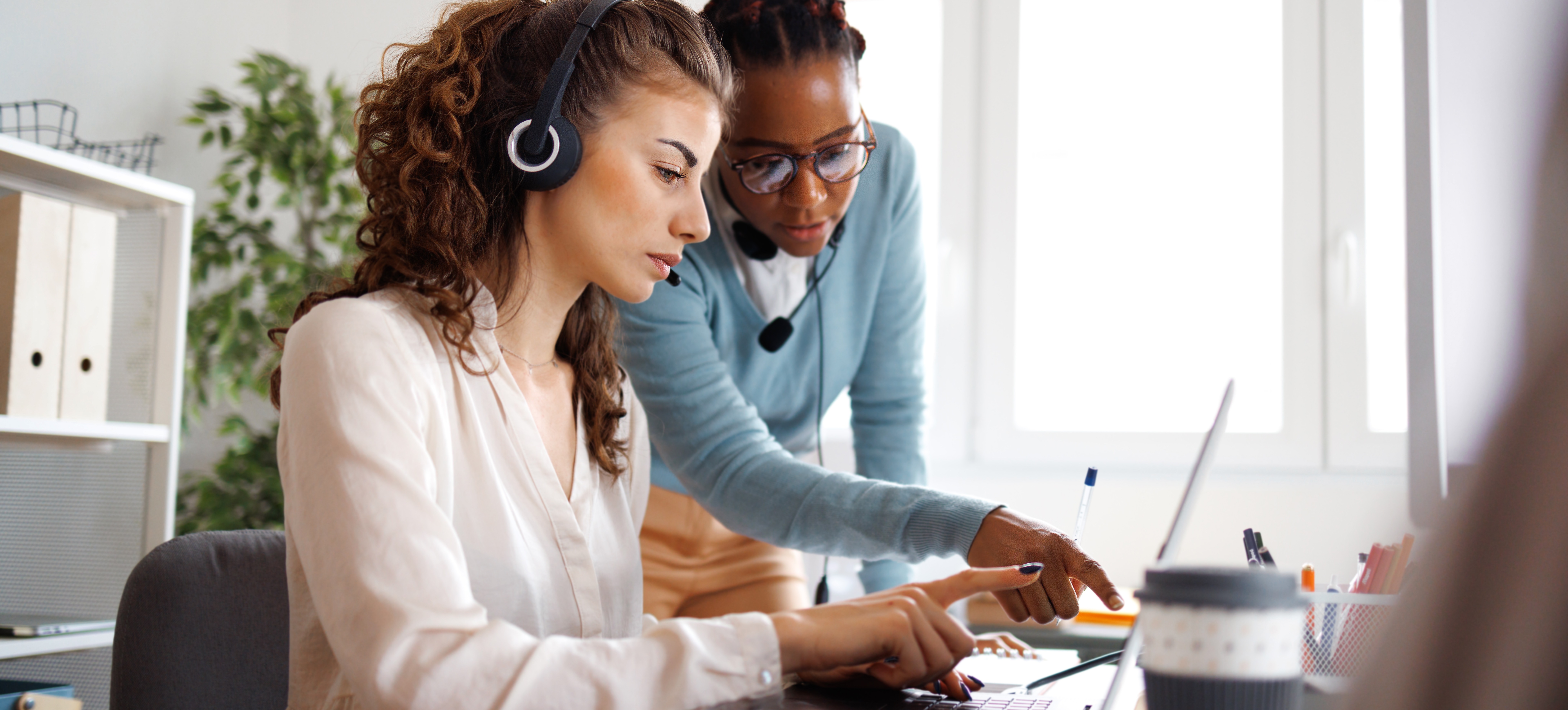 [Hauptbild] Eine Computer-Support-Spezialistin mit Headset steht über ihrem Kollegen und zeigt auf etwas auf dessen Laptop.