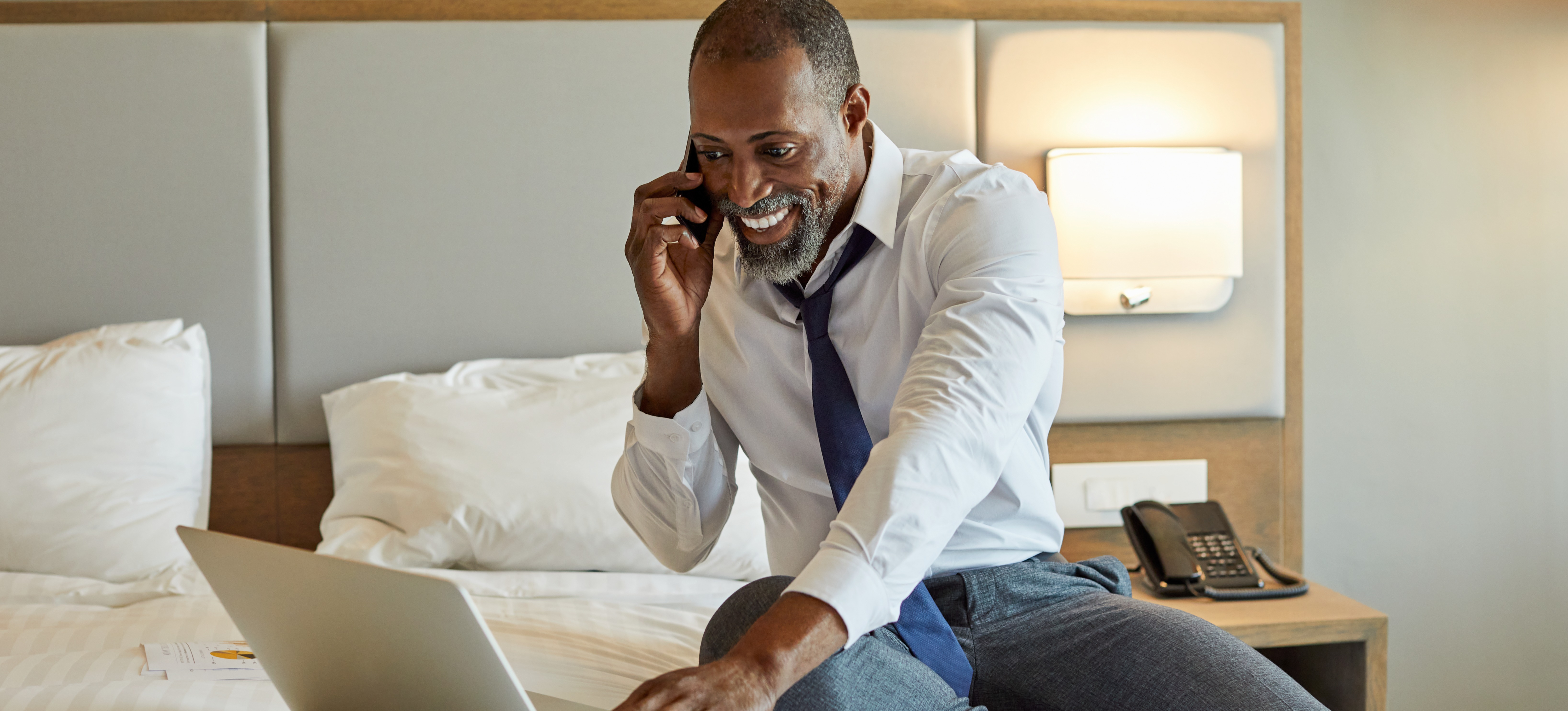[Featured Image] A person sits on a bed in a hotel room while using a smartphone and laptop to work on a project for their job in cybersecurity in the hospitality industry. 
