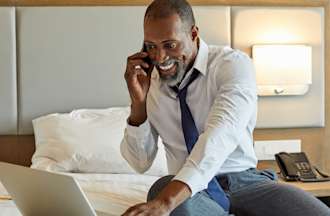 [Featured Image] A man sits on a bed in a hotel room while using a smartphone and laptop to work on a project for his job in cybersecurity in the hospitality industry. 
